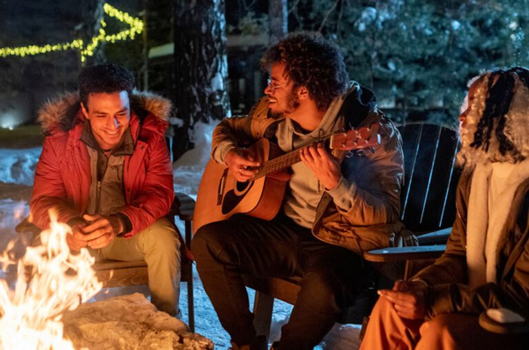 A group of people sitting outside by a firepit, one is playing a guitar. A group of people sitting outside by a firepit, one is playing a guitar.